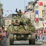 Reconstruction tank with re-enactors in uniform of American WWII cheered on by crowds in a street in Normandy