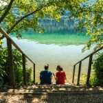 Man and woman sit looking over a crystal clear lake surrounded by forest in Isere, eastern France