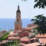 Pointed church tower reaching above terracotta roof tops, Mediterranean sea in the background at Menton, France